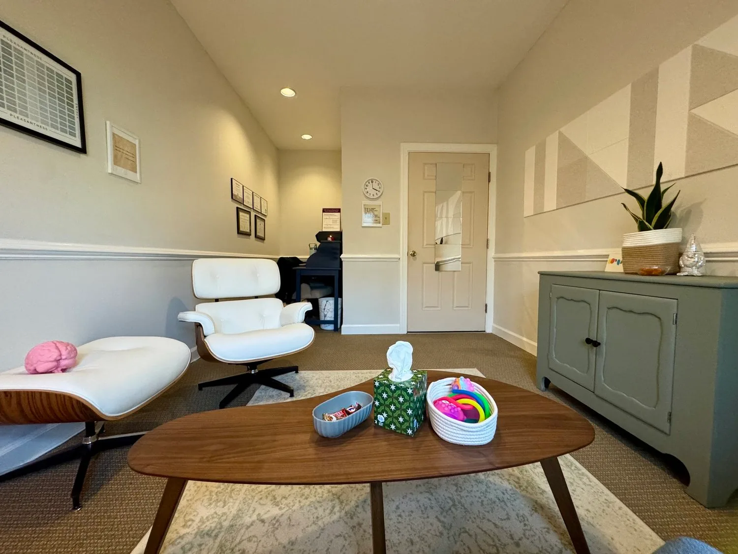 Indoor reception area with white modern lounge chair, a wooden table with toys, and a textured green sideboard with decorative items and a potted plant. The wall has framed artwork and a round clock. There is a door with a mirror and folder frames, and recessed ceiling lights.