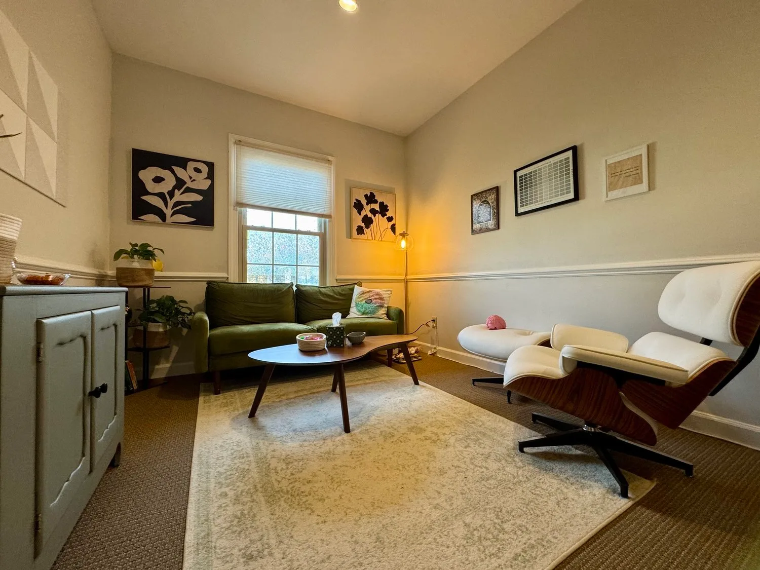 Living room with a green couch, a white armchair, a coffee table, wall art, a window with blinds, and a lamp in the corner.